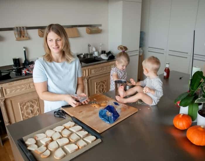 photo of mom making sandwhiches with her children sitting to her left on the counter playing with food