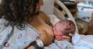 The image shows a mother in a hospital bed and hospital gown breastfeeding a newborn infant