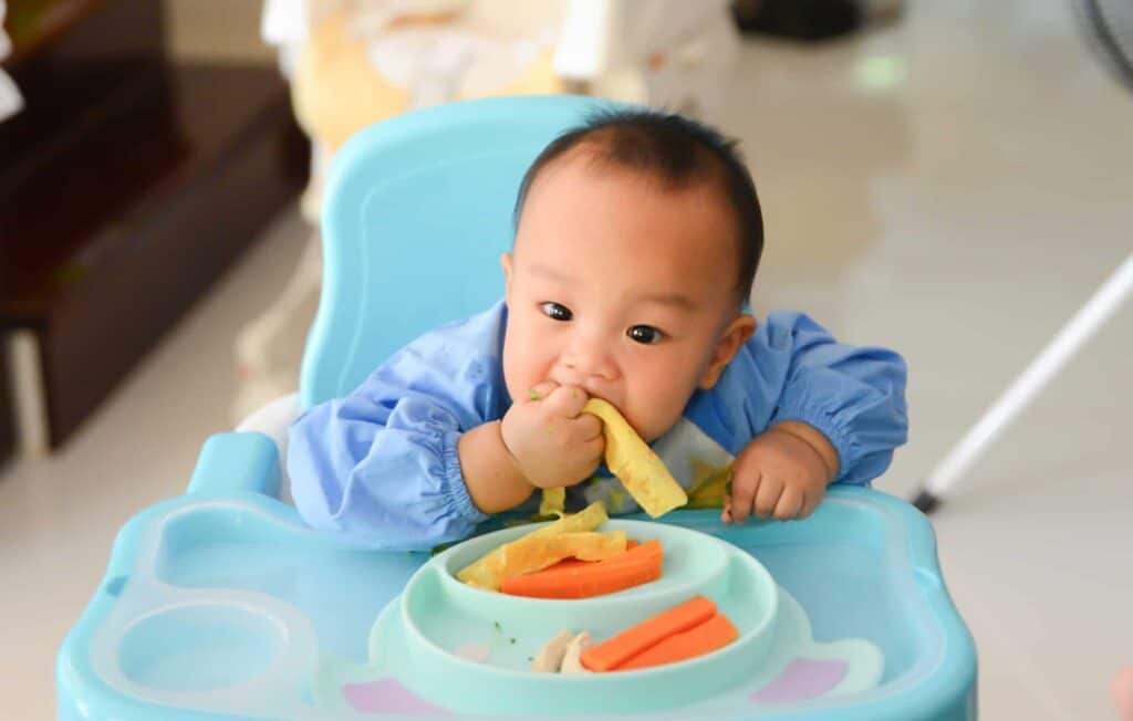 Baby boy bringing food to his mouth and eating while sitting in blue high chair