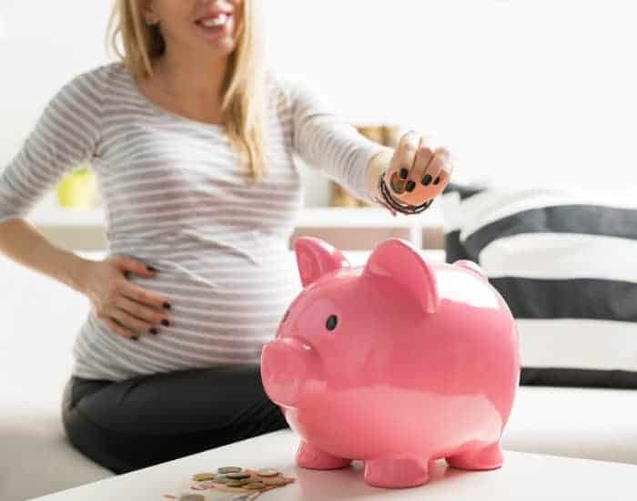 pregnant woman placing change (coins) into pink piggy bank (in article titled: how to afford newborn essentials on a budget)