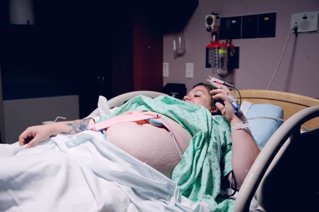 woman in labor with stretch marks wearing a green hospital gown, laying in a hospital bed, and hooked up to monitoring.