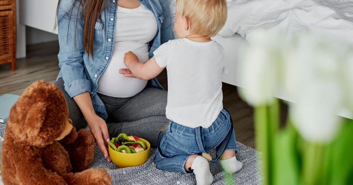 image of pregnant mother sitting on the ground next to her child who is touching her pregnant belly
