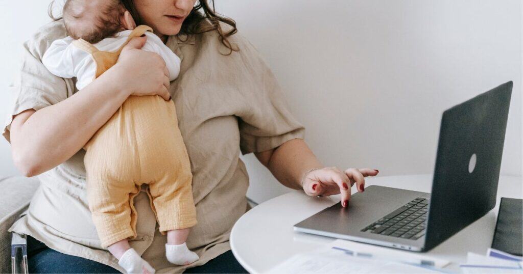 Woman holding a baby with a yellow outfit while using a laptop at a small table. Image from Canva.