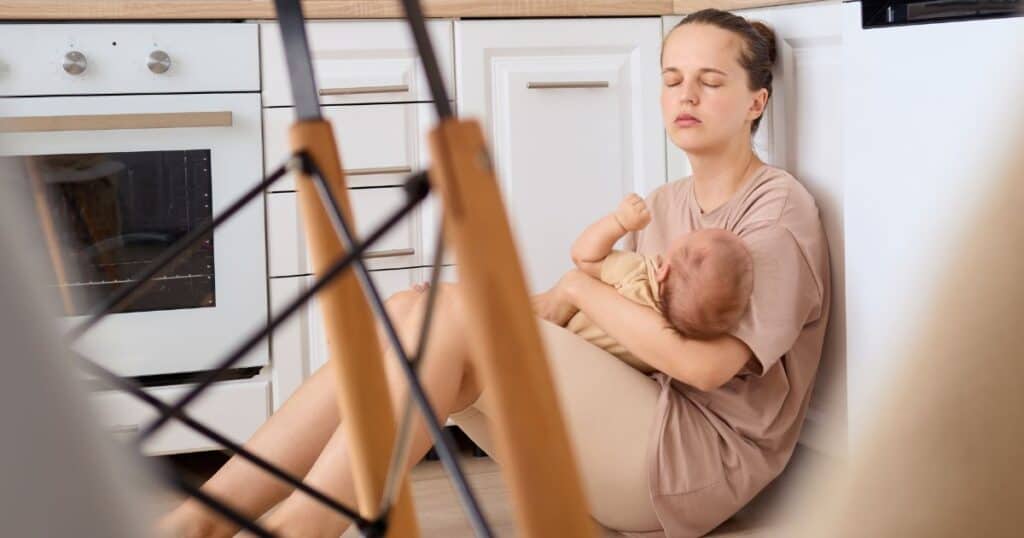 image of a white woman holding a newborn while sitting on a floor in a kitchen looking tired and exhausted