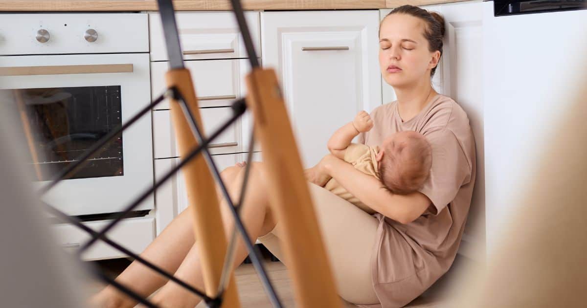 image of a white woman holding a newborn while sitting on a floor in a kitchen looking tired and exhausted