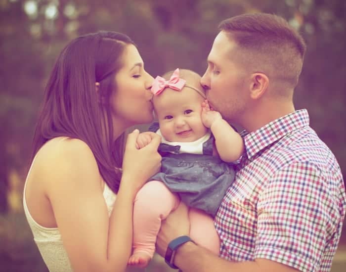 two parents (man and woman) holding their baby up between them and giving it a kiss on the cheek. 