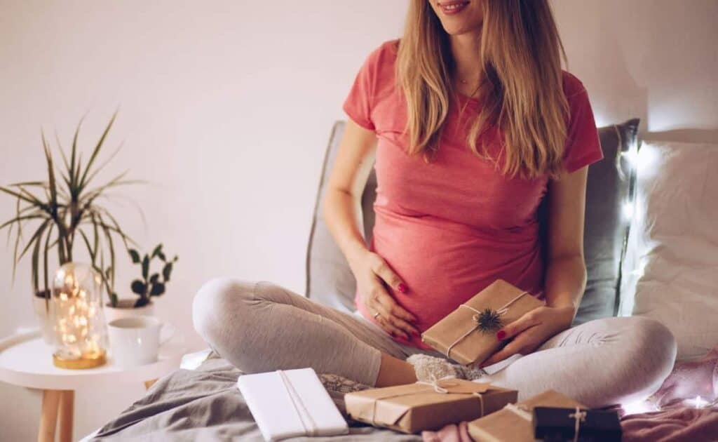 Image shows a pregnant woman in a red shirt sitting criss crossed on a bed, holding some gifts next to a nightstand