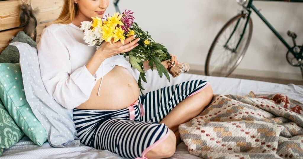 photo of pregnant woman holding flowers while sitting on a bed wearing black and white striped pants