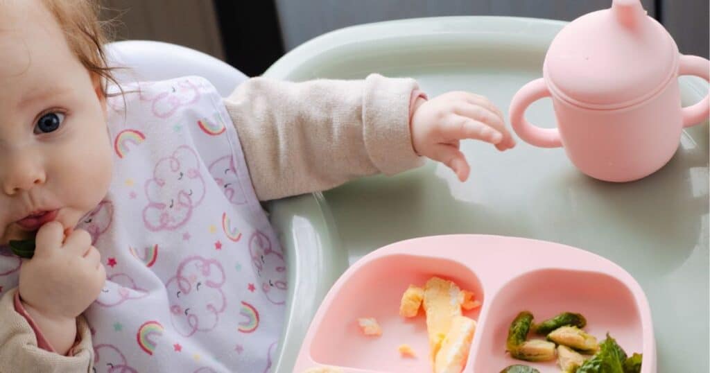 image of baby looking at the camera while sitting in a high chair eating food with pink plate and cup.