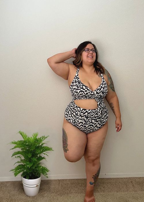 image of myself (a woman with brunette hair, glasses, and tattoos) standing next to an indoor plant, wearing a Knix wrap swimsuit in a black/white color