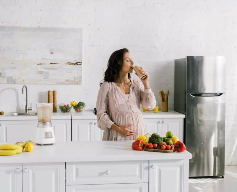 Pregnant woman standing in white kitchen with one hand on her belly and the other drinking a protein shake (image within the article titled:Is Using Protein Powder While Breastfeeding Safe?)