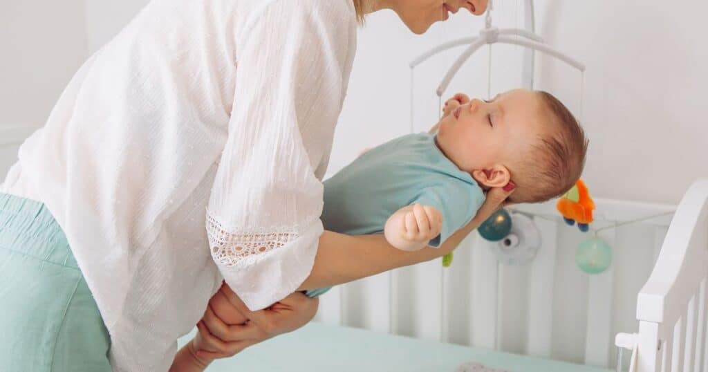A mother gently lowering her baby into a crib. The baby, dressed in a light blue onesie, is cradled in the mother's arms, appearing peaceful and calm. A mobile with colorful hanging toys is visible above the crib, adding a soft, playful element to the nursery scene.