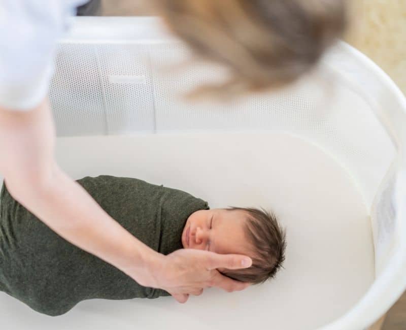 Infant baby swaddled inside of a crib as woman reaches down touching it's cheek.