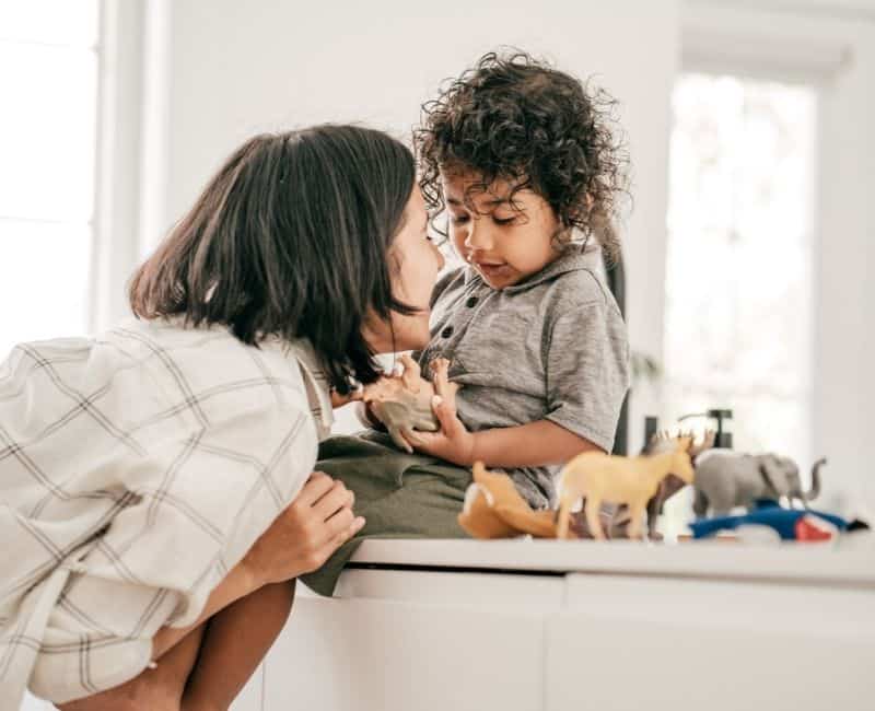 picture shows a young child with curly hair sitting on a counter with woman smiling scrunched below looking up at him. 