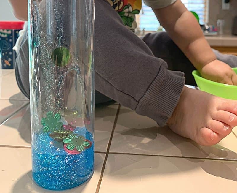 picture of a glitter glue sensory bottle in the making. Bottle is clear and has blue glitter glue and some beads and sequins inside. bottle is on the counter next to a young child's foot