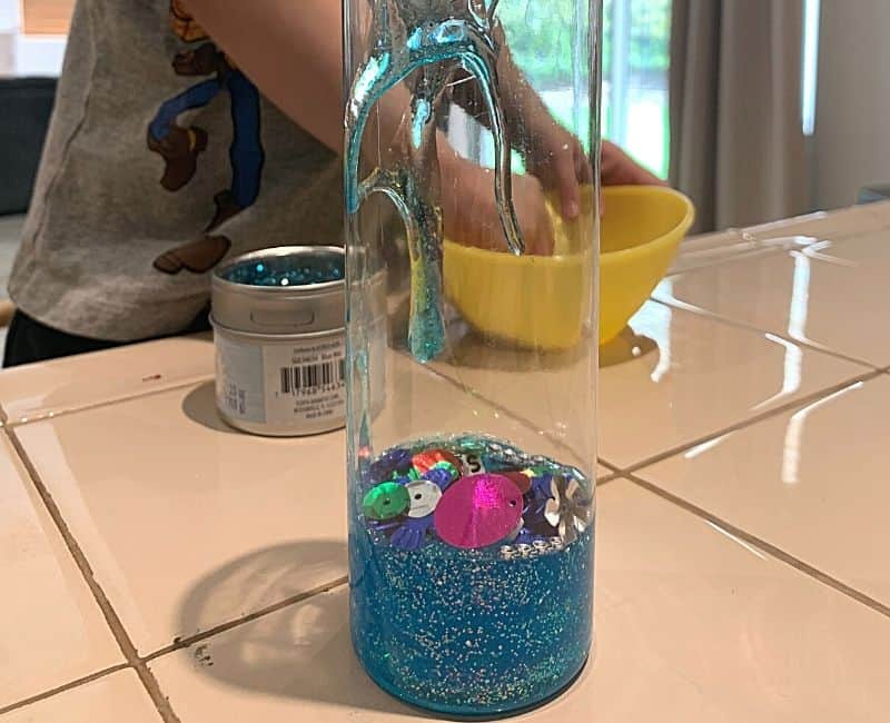 picture of a glitter glue sensory bottle in the making. Bottle is clear and has blue glitter glue and some beads and sequins inside.