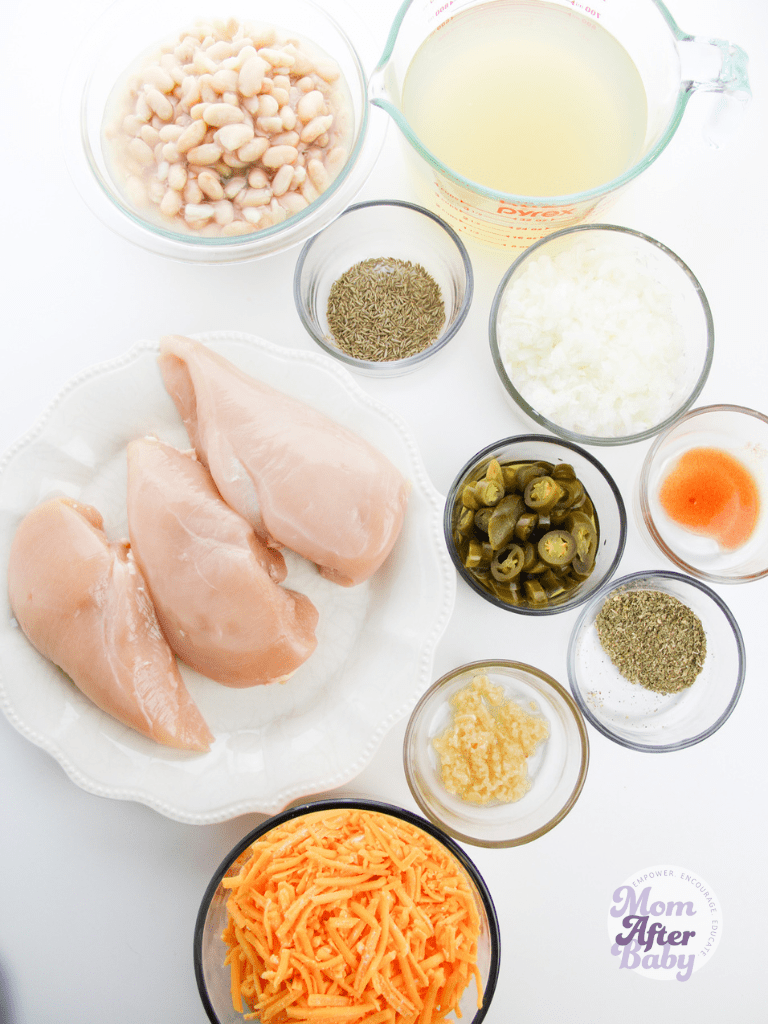 Flat lay photo of white chicken chili ingredients: raw chicken breasts, bowl of cheddar cheese, jalapenos, oregano, garlic, and lots of spices