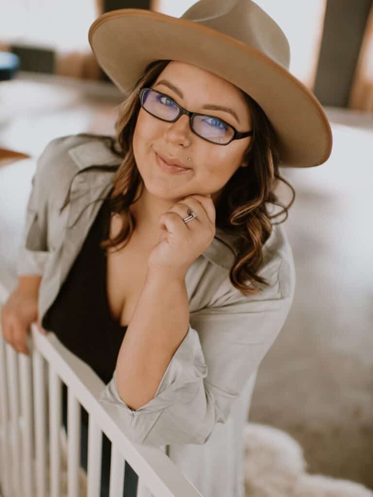 headshot image of Karissa from Mom After Baby leaning on a baby crib, smiling at the camera and wearing a hat