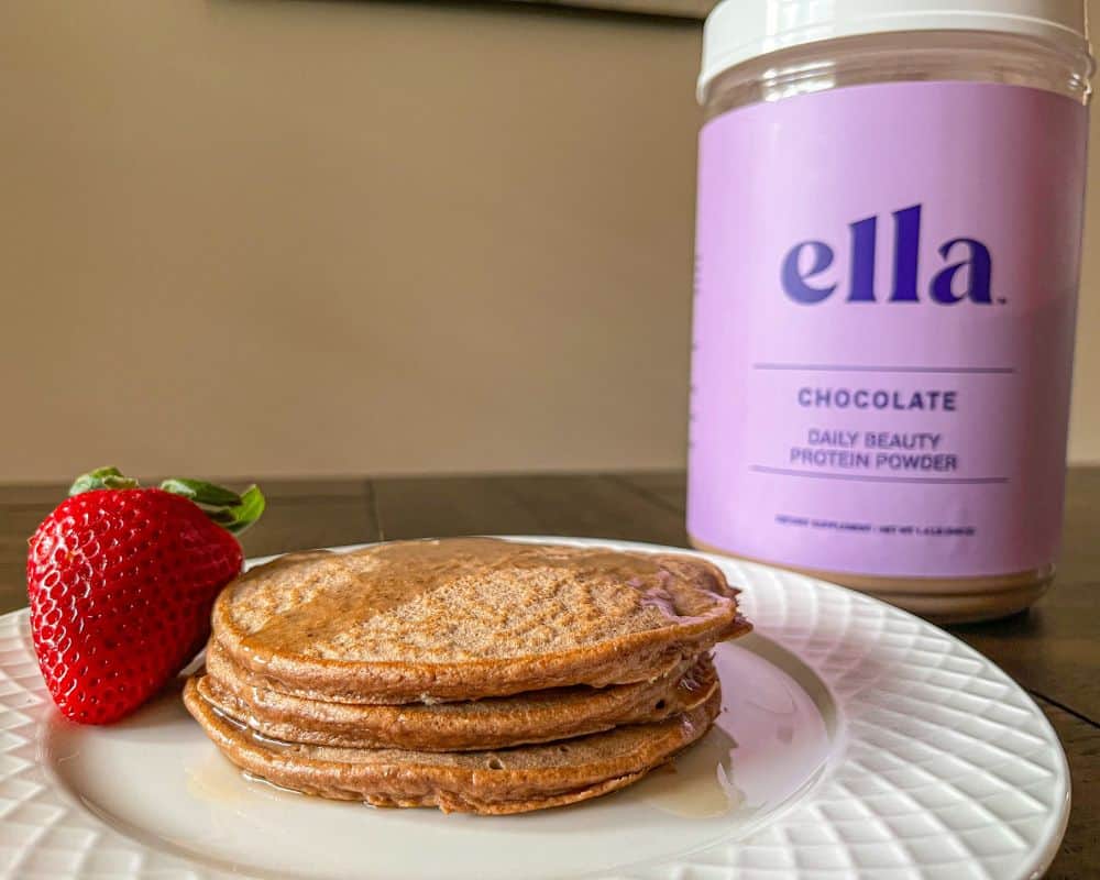 photo of the cooked ella beauty protein powder pancakes on a white plate, next to a strawberry and next to the container of ella collagen protein