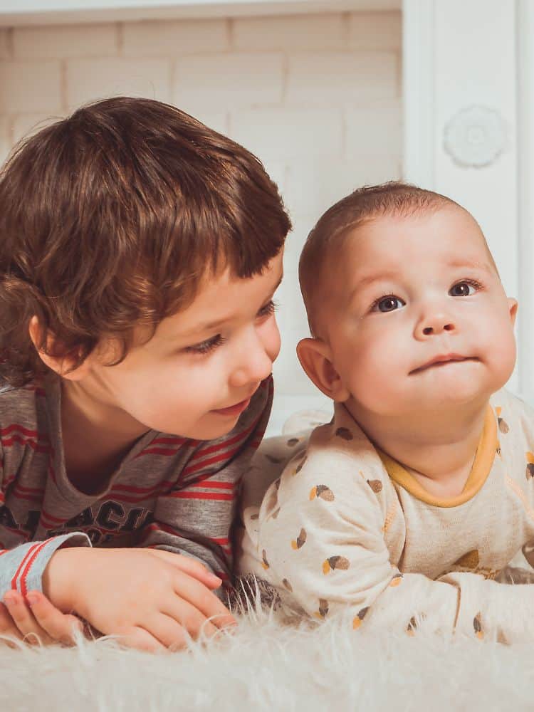 image shows a toddler boy with brown hair laying on his stomach and looking over at a baby sibling thats looking up at the camera