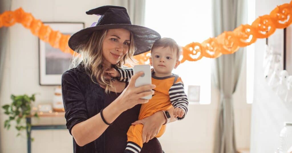 This image shows a woman wearing black and a black with hat holding a baby dressed as a pumpkin, smiling while taking a selfie in a mirror.
