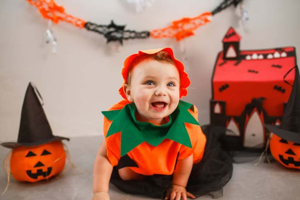 this image shows a baby smiling and happy, dressed as a pumpkin. In the background there is a pumpkin with a hat and some halloween decor hanging from the wall.