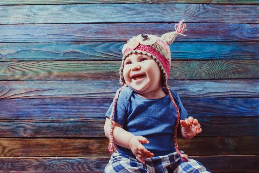 Image shows a smiling baby in a blue shirt wearing and owl knit beanie. Background is a colorful blue/brown paneled wall.