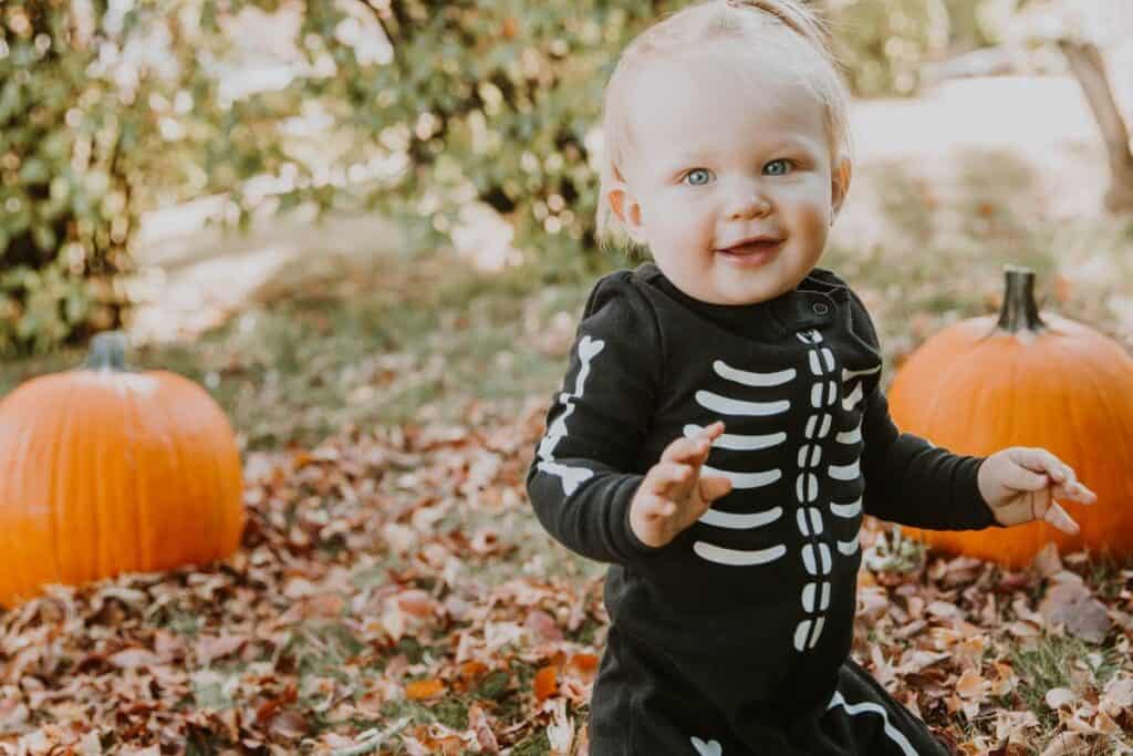 This image shows an older baby outside standing in fall leaves wearing a black and white skeleton onesie. There are some pumpkins in the background alslo.