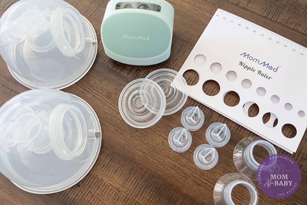 This image shows the Mommed breast milk collection cups, accessory parts, and a nipple measuring guide on top of a brown table