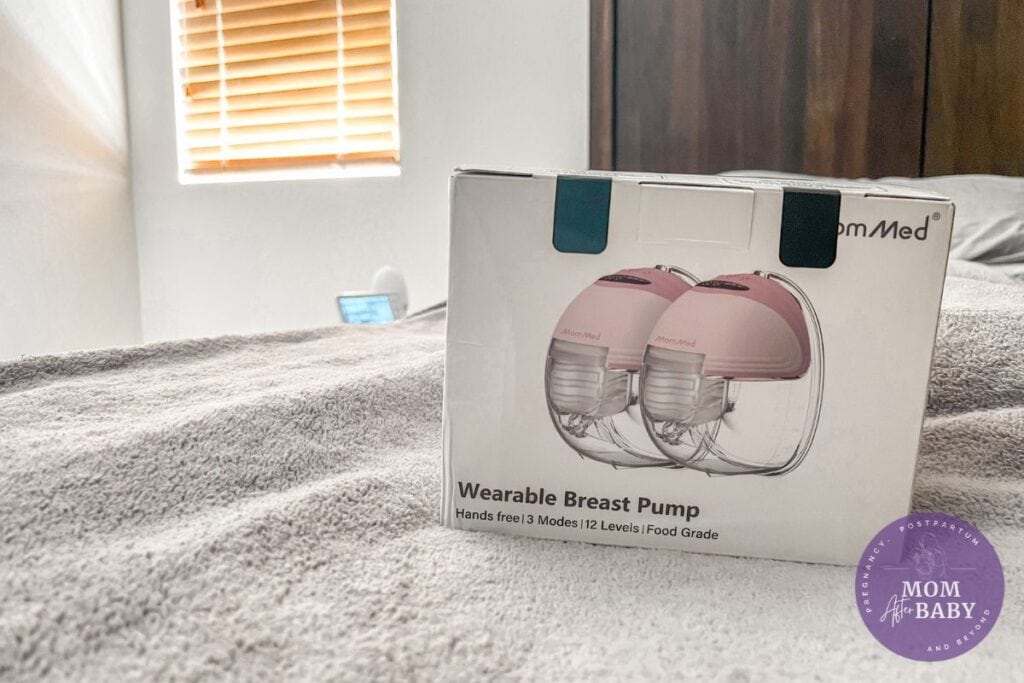 Image shows the MomMed Wearable Breast Pump box on my bed (on top of a grey blanket) with dark wood headboard in the background and my son's baby monitor on the nightstand