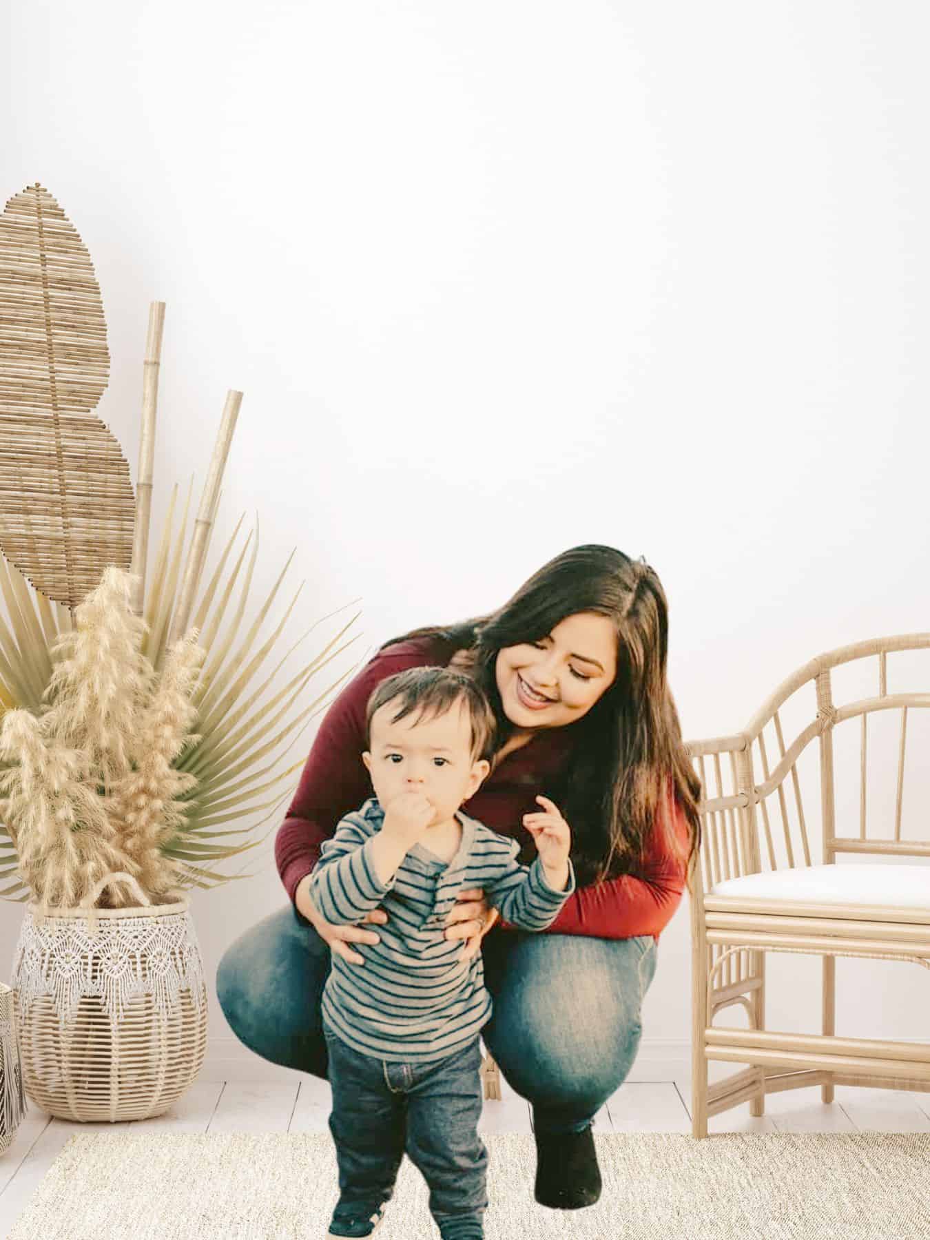 this image shows me wearing a read shirt, with dark brown hair, kneeling down to hold and look at my son who is eating a cookie, and wearing blue. The background shows a boho styled room