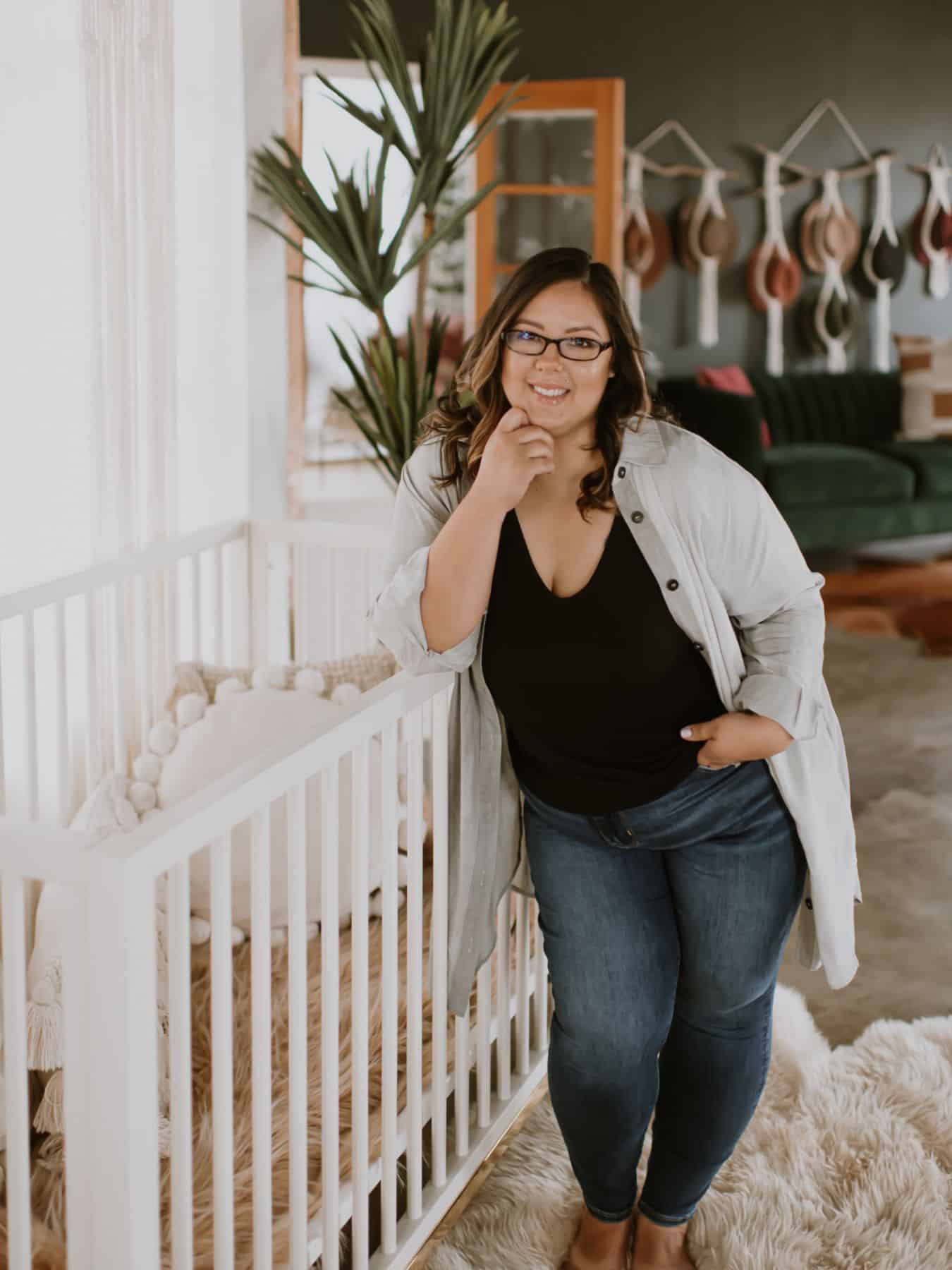 this image shows karissa of mom after baby standing and leaning on a white baby crib with her hand under her chin, smiling at the camera.