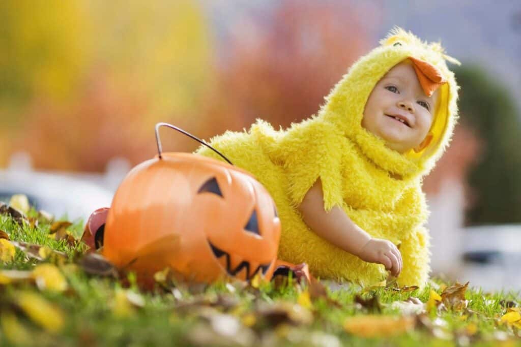 This image shows a baby sitting next to a jack-o-lantern candy bucket in the grass smiling, while wearing a yellow chicken costume