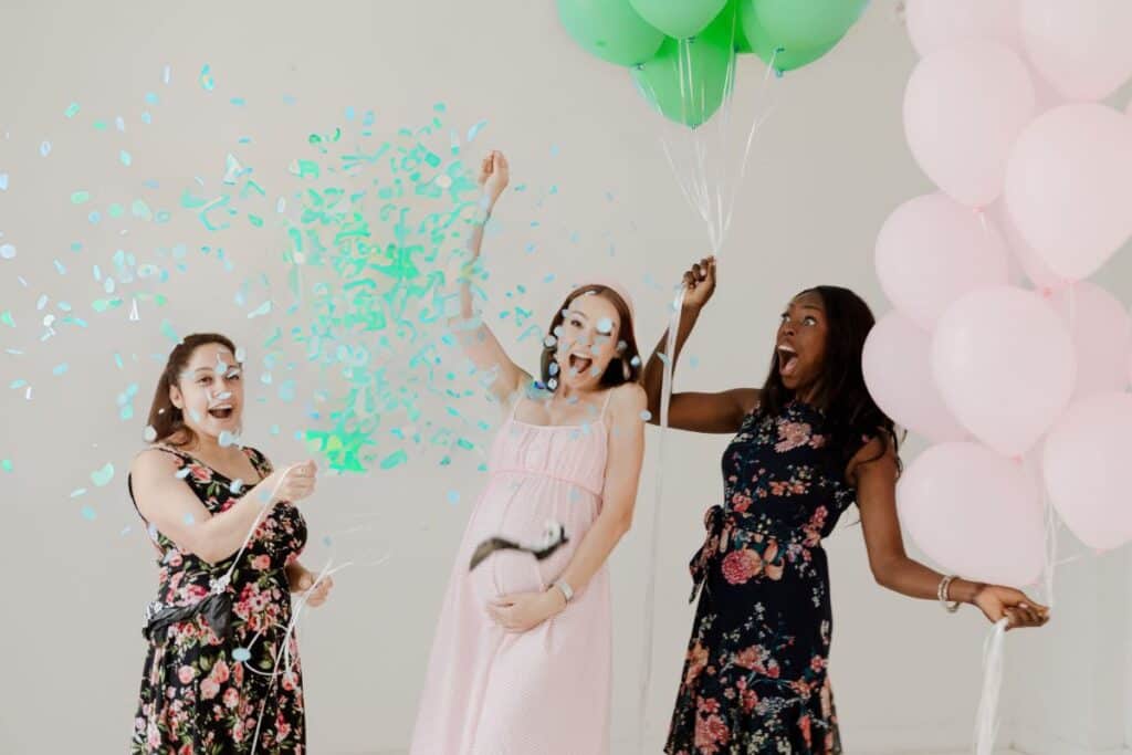 Image shows a pregnant woman popping a green gender reveal balloon alongside 2 other women celebrating with her