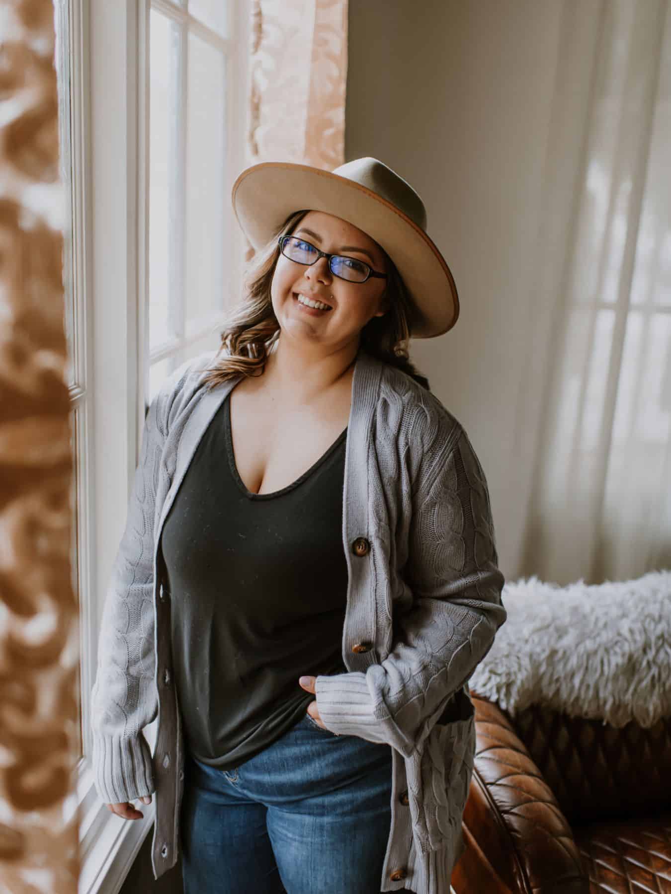 image shows karissa of mom after baby standing facing a window, wearing a hat, with her hand in her pocket while looking at the camera and smiling