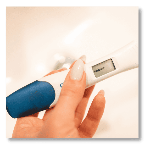 image shows a woman's hand with long nails holding a positive pregnancy test with a blue cap.