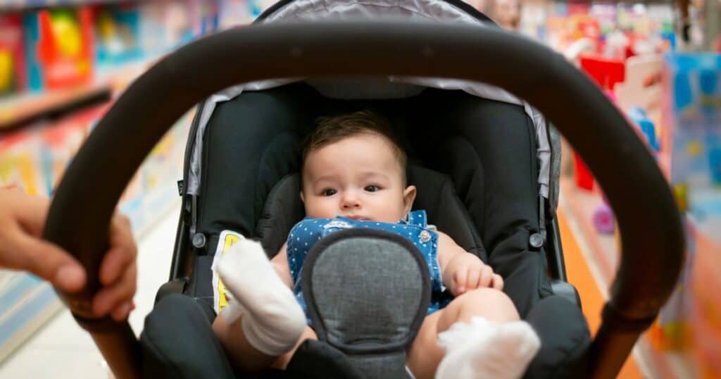 image shows a close up of an infant sitting inside of a travel stroller with a blurred out, colorful background that appears to be a store of some kind.