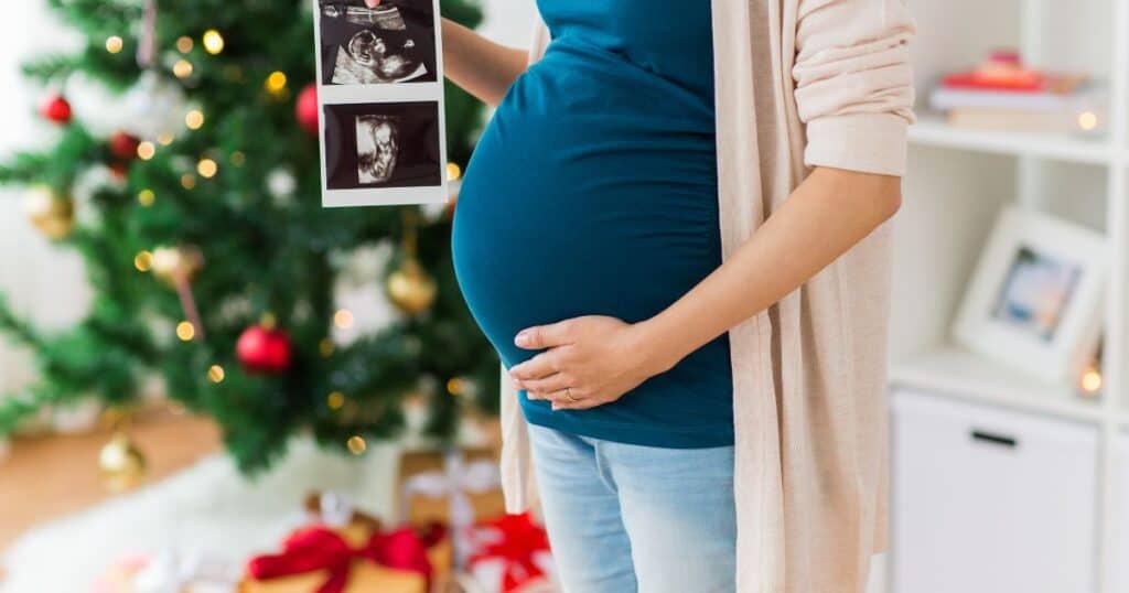 New Year Baby Announcement Ideas cover image of a woman in a teal shirt holding her belly with an ultrasound photo of a baby next to a christmas tree
