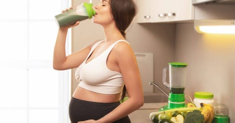 image of a pregnant woman standing in a kitchen drinking a shake that appears to be green in color