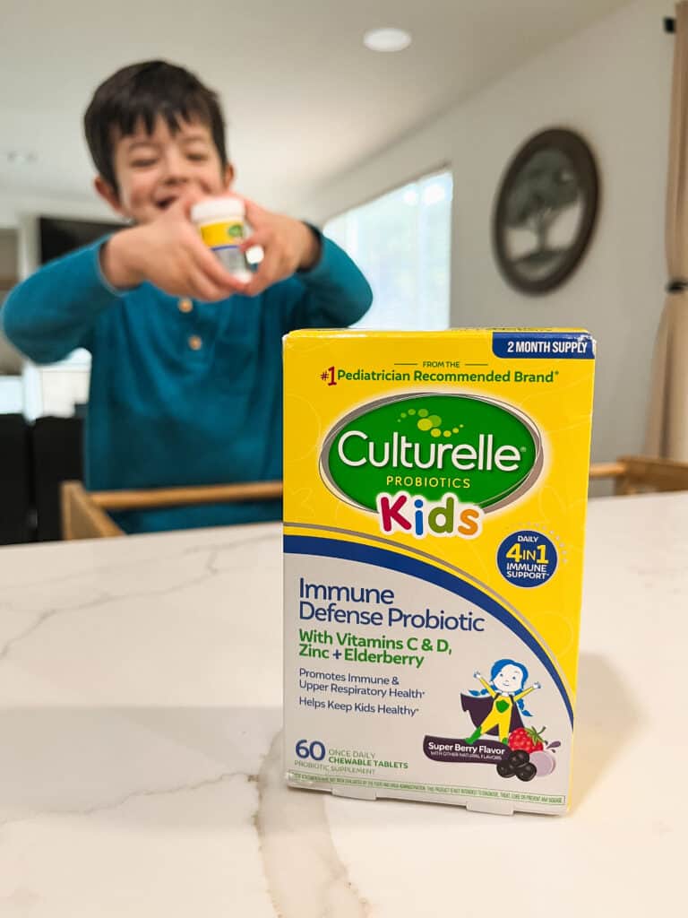 Toddler happy and holding his Culturelle Probiotic bottle with a yellow Culturelle probiotic box on the counter infront