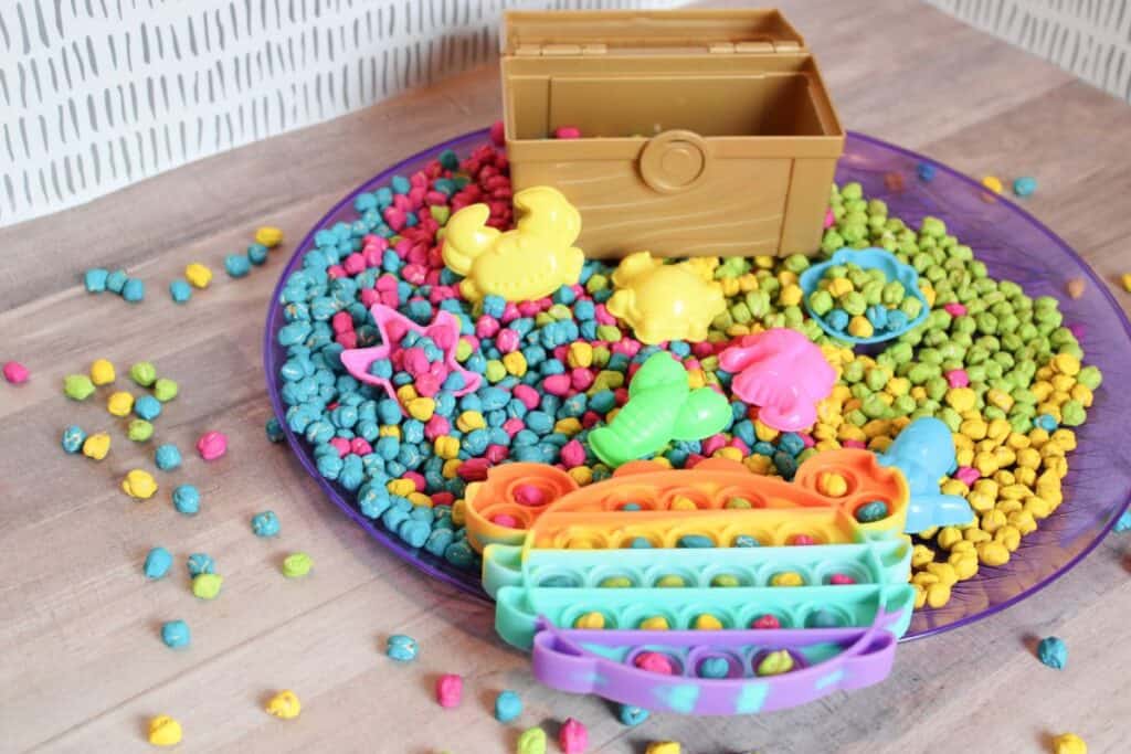 Image of a chickpea sensory bin put together on a large purple tray with various toys and a small treasure chest.
