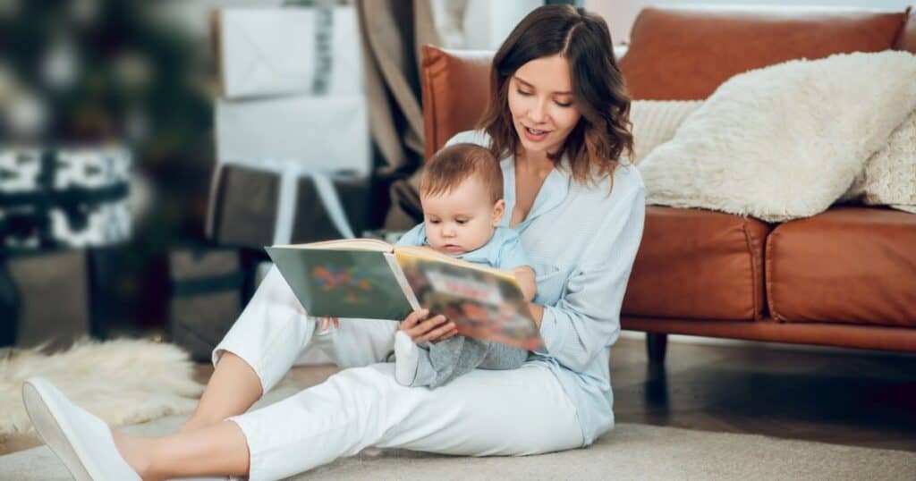 image shows a brunette haired woman sitting down, holding a baby in her lap while reading a book to baby. she's sitting up against a brownish/red sofa.