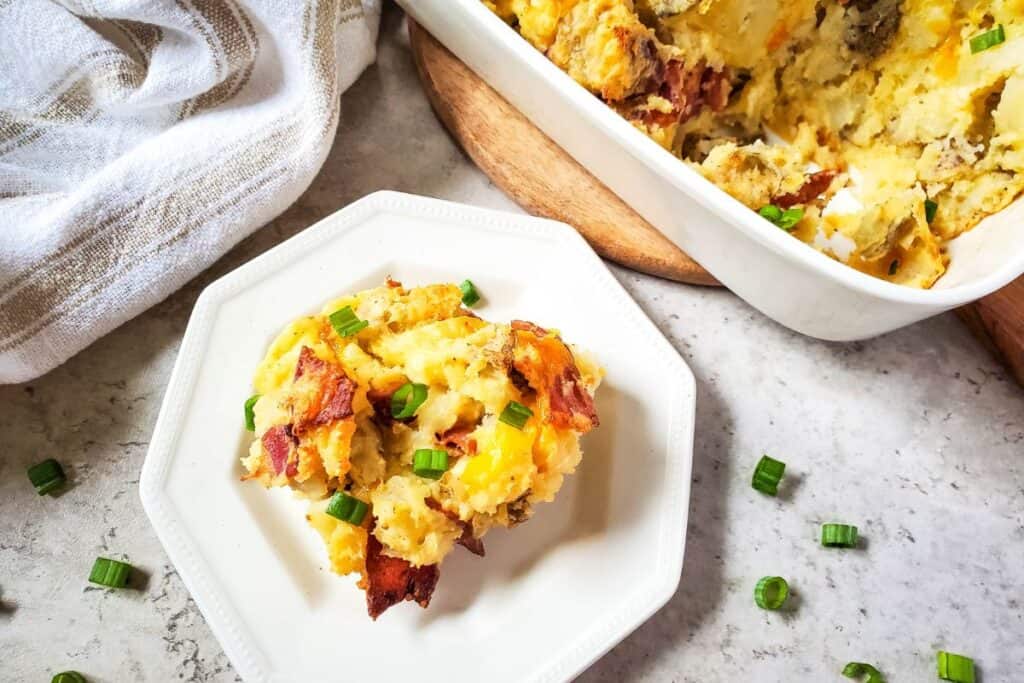 Twice baked potato casserole finished dish presented nicely on a white plate next to the baking dish and a dish cloth