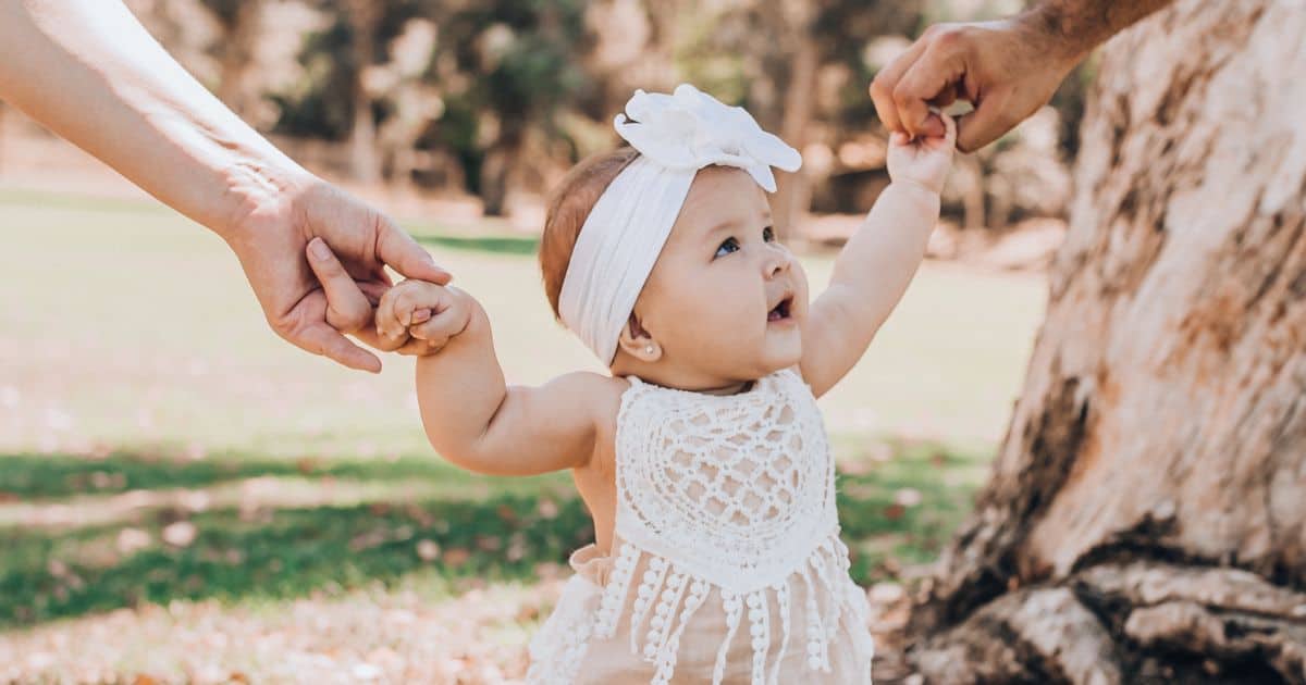 close up image of a baby girl holding a parent's hand on each side while looking up at one of them and wearing a white headband