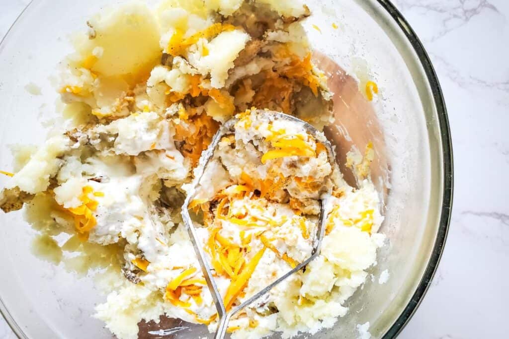 image shows cooked potato along with other ingredients being mashed and mixed with a handheld potato masher