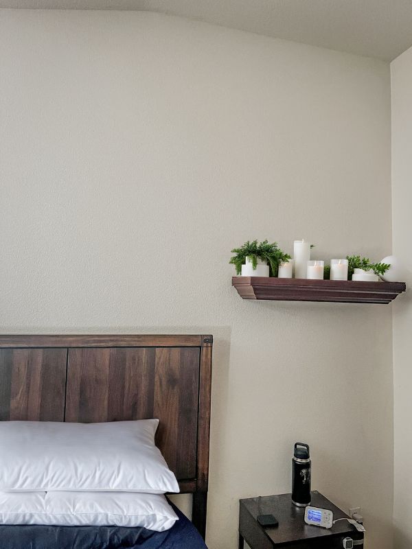 image of the corner of a headboard with grey pillows, a floating shelf on the top right and a brown wooden dresser at the bottom.