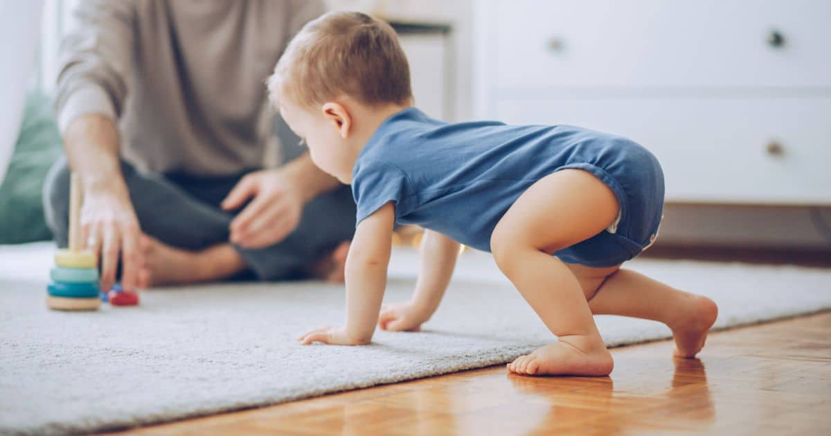 baby in a blue onesie crawling on the ground, onto a light colored carpet towards parent in a green shirt.