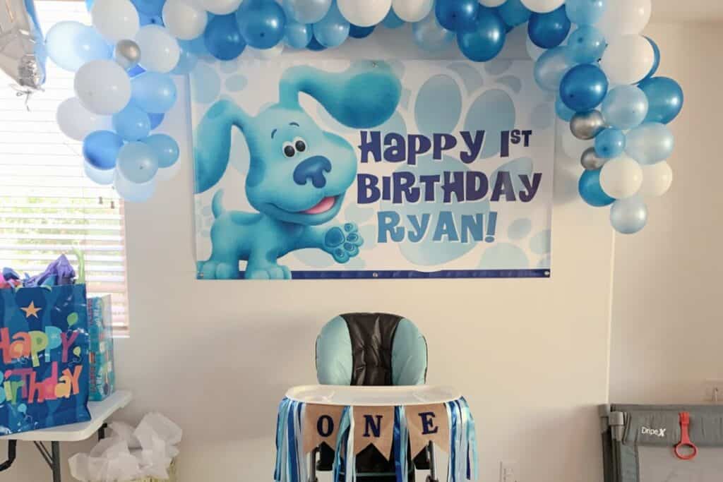 image shows a decorated background for baby's first birthday with a blue and while ballooon arch, a happy 1st birthday blue's clues sign, and a highchair with a ONE banner.