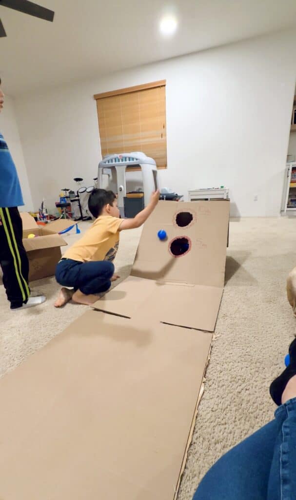 image of a young child in an orange shirt playing skeeball made from a cardboard box in living room
