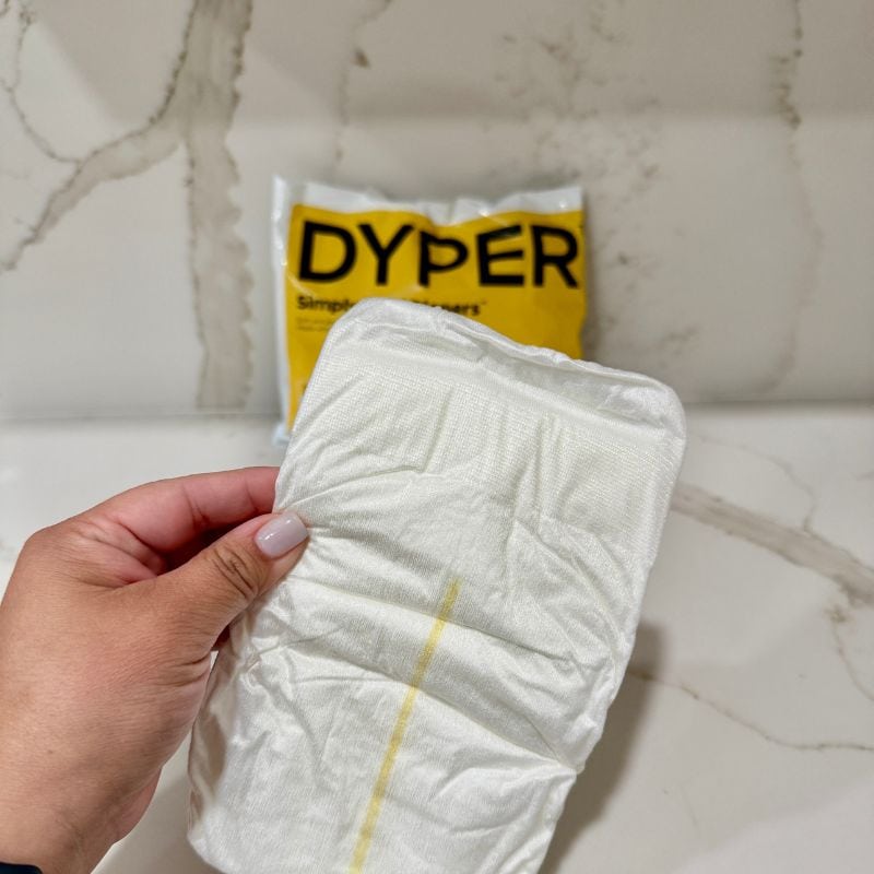 close up image of my hand holding the Dyper baby Diaper with the Dyper yellow packaging in the background